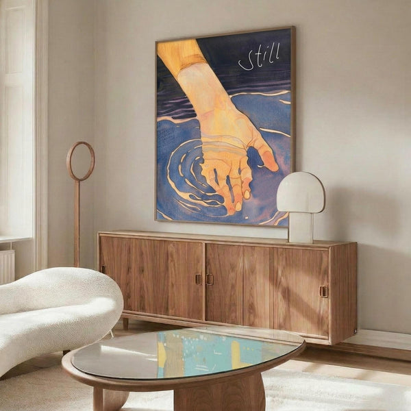 May include: Modern living room with walnut credenza displaying framed poster showing golden hand touching blue water with ripples, creating concentric circles. White sculptural chair and oval glass coffee table in foreground. Warm wood tones complement blue and orange artwork colors. Contemporary minimalist interior styling with natural lighting.