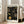 May include: Framed poster with black background featuring golden yellow circular dots and spiral patterns in Kusama's signature polka dot style. White text reads "YAYOI KUSAMA" at top and "Modern Art 1954" at bottom. Light wood frame leans against white wall next to beige upholstered chair in bright, naturally lit room with hardwood floors and large windows.