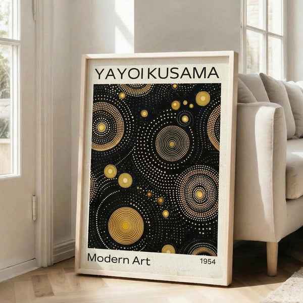 May include: Framed poster with black background featuring golden yellow circular dots and spiral patterns in Kusama's signature polka dot style. White text reads "YAYOI KUSAMA" at top and "Modern Art 1954" at bottom. Light wood frame leans against white wall next to beige upholstered chair in bright, naturally lit room with hardwood floors and large windows.