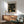 May include: Framed art print featuring black background with golden yellow dotted circular patterns in varying sizes, hung above wooden sideboard with table lamp. Mid-century modern room setting with sage green upholstered chair, round coffee table, and natural sunlight casting shadows on white wall.