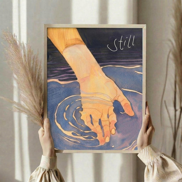May include: Person holding framed artwork showing hand touching water creating ripples, with word "still" in white text. Watercolor style illustration in blue, orange and yellow tones. Light wood frame, neutral beige clothing, dried pampas grass visible, soft natural lighting against white curtained background.