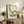 May include: Framed vintage-style poster featuring green mid-century modern lounge chair illustration on cream background, displayed on rustic wooden credenza alongside fern plant, ceramic vases, and books in neutral-toned living room setting