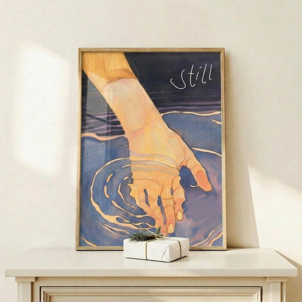 May include: Framed poster showing a hand reaching down to touch water, creating ripples in blue and purple tones. The word "still" appears in white text at the top. The artwork has a dreamy, watercolor-like quality with warm peachy skin tones against cool blue water. Displayed on a light wood surface with a small wrapped gift box nearby, set against a white wall.