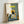 May include: Framed art print leaning against white wall showing black cat sitting at kitchen table with yellow "CHOW!" text above. Scene depicts breakfast spread with cookies, eggs, orange juice, and coffee on blue checkered tablecloth. Wooden chairs visible, artwork has painterly illustration style with sage green and cream background. Light wood frame surrounds the print.