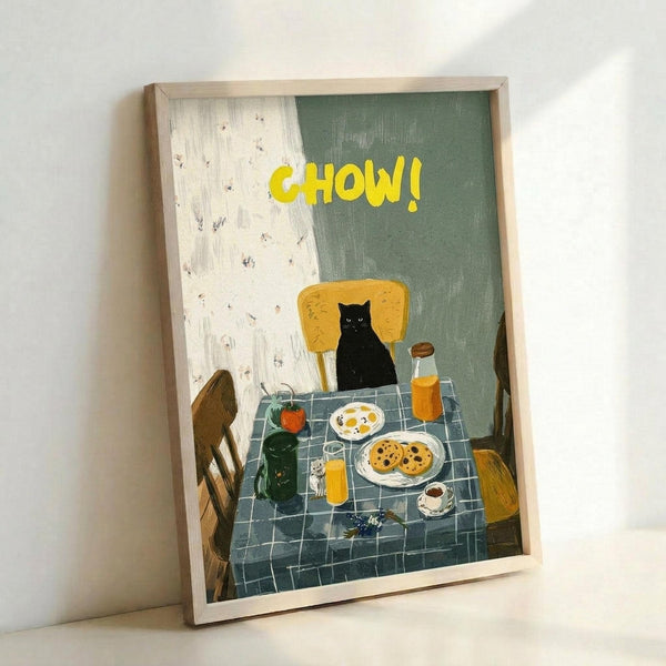May include: Framed art print leaning against white wall showing black cat sitting at kitchen table with yellow "CHOW!" text above. Scene depicts breakfast spread with cookies, eggs, orange juice, and coffee on blue checkered tablecloth. Wooden chairs visible, artwork has painterly illustration style with sage green and cream background. Light wood frame surrounds the print.