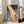 May include: Framed art print leaning against white wall beside beige sofa in bright living room with large windows. Artwork shows hand touching blue water creating ripples, with golden yellow skin tones against deep blue water and dark sky. White cursive text reads "still" in upper right corner. Light wood frame, approximately 2-3 feet tall.