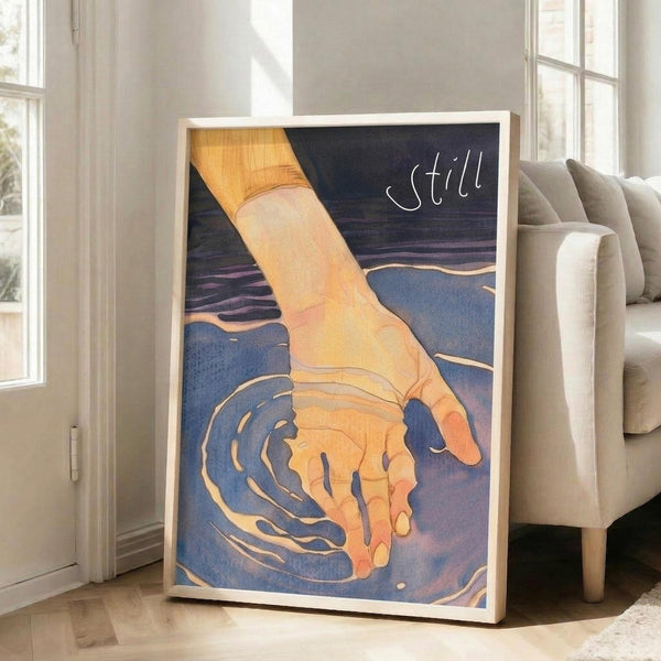 May include: Framed art print leaning against white wall beside beige sofa in bright living room with large windows. Artwork shows hand touching blue water creating ripples, with golden yellow skin tones against deep blue water and dark sky. White cursive text reads "still" in upper right corner. Light wood frame, approximately 2-3 feet tall.
