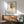 May include: Framed abstract art print featuring geometric shapes in yellow, blue, red, and green with black linear elements on cream background, displayed above mid-century modern wooden credenza with table lamp, next to sage green upholstered armchair with wooden frame in sunlit room with white walls