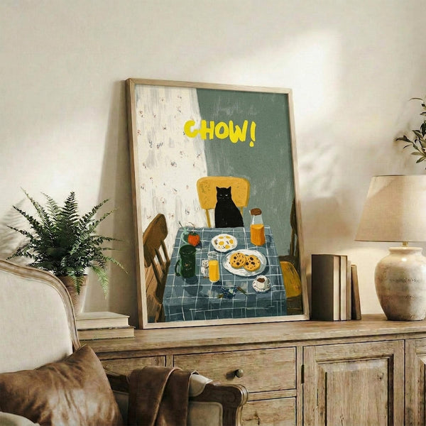 May include: Framed poster displaying a whimsical painted scene of a black cat sitting at a breakfast table with yellow "CHOW!" text above. The artwork shows a checkered blue tablecloth, plates with eggs and cookies, orange juice, and wooden chairs in folk art style. Frame sits on rustic wooden sideboard alongside fern plant, books, and ceramic vases in neutral-toned living room setting.