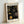 May include: Framed art print leaning against white wall showing abstract pattern of golden yellow circles and dots on black background, displayed in light wood frame on light wooden surface