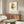 May include: Abstract geometric art print featuring colorful overlapping rectangles and linear elements in yellow, blue, red, and black on cream background, displayed in thin wooden frame above mid-century modern walnut credenza. Room scene shows white sculptural chair, brass floor lamp, oval glass coffee table, and neutral walls with natural lighting creating soft shadows.