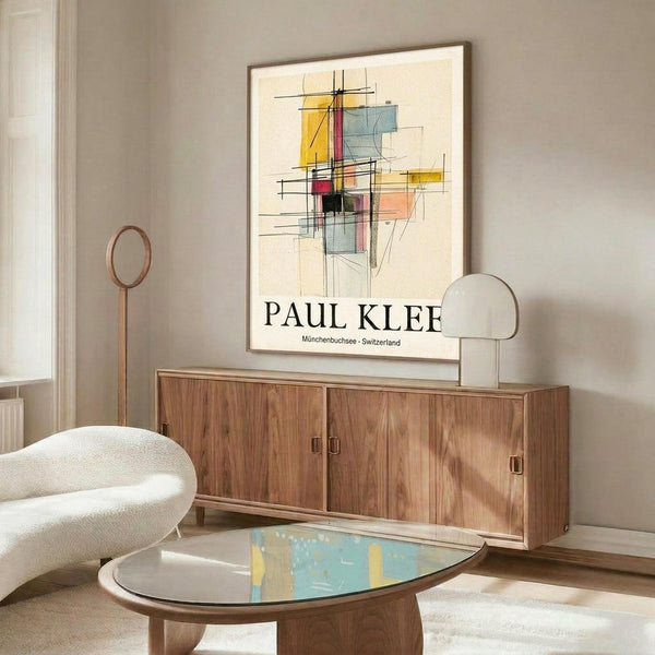 May include: Abstract geometric art print featuring colorful overlapping rectangles and linear elements in yellow, blue, red, and black on cream background, displayed in thin wooden frame above mid-century modern walnut credenza. Room scene shows white sculptural chair, brass floor lamp, oval glass coffee table, and neutral walls with natural lighting creating soft shadows.