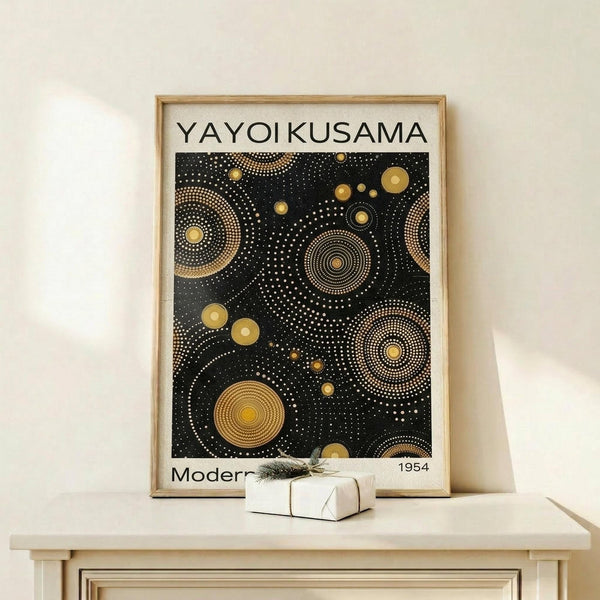 May include: Framed poster print with black background and golden circular dot patterns in concentric rings, displayed on light wood shelf against white wall. Small white wrapped gift box with greenery sits beside the wooden frame. Abstract geometric design features various sized circles and dotted spiral motifs in gold and yellow tones.