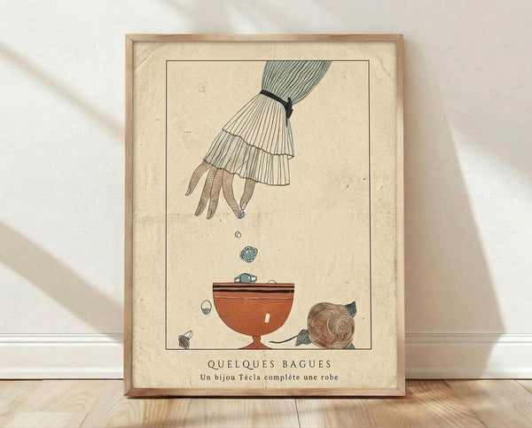 May include: Framed vintage French fashion illustration showing an elegant hand with ruffled sleeve dropping jewelry into an ornate bowl. Art features muted earth tones on cream textured paper with French text below. Natural wood frame sits on light wooden floor against white wall with soft natural lighting creating gentle shadows.