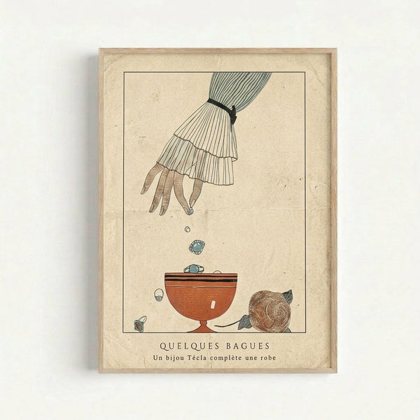 A vintage French art print in a light wood frame showing an elegant hand with ruffled sleeve cuff dropping jewelry into an orange ceramic bowl. The illustration features delicate line work in muted colors on aged cream paper, with scattered rings and a seashell around the bowl. French text appears at the bottom of the print.