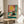 May include: Framed poster print displayed on wooden side table next to beige upholstered chair and plant. Artwork shows black cat wearing yellow sunglasses sitting poolside with turquoise tiles, coral pink wall, yellow sun, and tropical drink with straw. Natural lighting creates soft shadows across white wall.