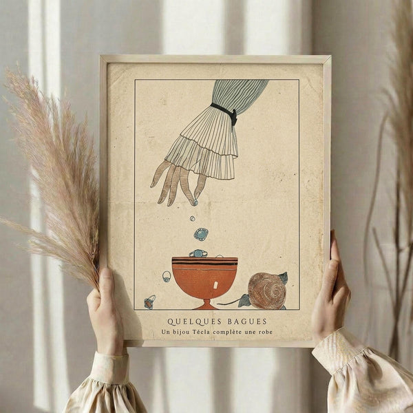 May include: Person in cream ruffled blouse holding framed vintage-style poster featuring an illustrated hand dropping jewelry into an ornate bowl, with decorative elements and French text below. Dried pampas grass visible on left side. Neutral beige wall background with soft natural lighting.