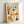 May include: Framed art print leaning against white wall showing twelve colorful illustrated portrait squares arranged in a grid. Each portrait features a different person with varied hairstyles, clothing, and facial features in warm tones of orange, yellow, green, and coral. The text "SOIS UNIQUE" appears at the top in black lettering. The artwork is displayed in a light wood frame on what appears to be a white surface.