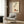 May include: Vintage 1920s Art Deco poster featuring an elegant hand with layered white sleeve cuffs dropping objects into a terracotta bowl, displayed in light wood frame on dark wood paneled wall above cream upholstered armchair with geometric patterned pillow, brass floor lamp and side table nearby