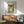 May include: Japanese-style woodblock print artwork hanging in modern living room with wooden credenza below, featuring mountain landscape with stream, cherry blossoms, and Japanese text. Print shows green hills, flowing water over stones, blooming trees under pale sky with moon. Room has mid-century teal armchair, white ceramic lamp, and warm natural lighting creating shadows on white wall.
