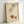 May include: Vintage-style art print in light wood frame leaning against white wall. Illustration shows hand in pleated sleeve dropping colorful marbles or gems into orange ceramic bowl, with decorative snail nearby. Cream textured background with French text at bottom. Whimsical early 20th century aesthetic with muted earth tones and delicate line work.