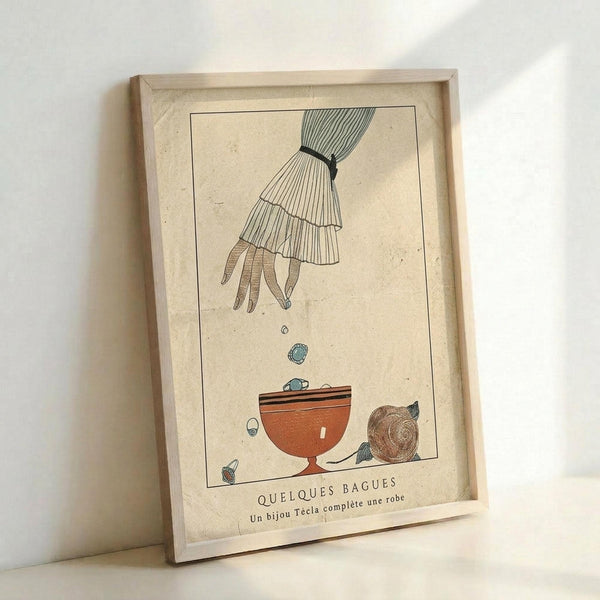 May include: Vintage-style art print in light wood frame leaning against white wall. Illustration shows hand in pleated sleeve dropping colorful marbles or gems into orange ceramic bowl, with decorative snail nearby. Cream textured background with French text at bottom. Whimsical early 20th century aesthetic with muted earth tones and delicate line work.