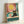 May include: Framed poster print leaning against white wall showing retro-style illustration of black cat wearing yellow sunglasses sitting by turquoise swimming pool. Cat positioned on beige tile pool edge next to pink drink with striped straw and lemon slice. Background features coral pink wall with large yellow sun and vintage-style faucet. Artwork has warm vintage color palette in natural wood frame.