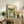 May include: A framed Japanese woodblock-style poster displaying a serene mountain landscape with flowing stream, cherry blossoms, and traditional text characters, positioned on a rustic wooden sideboard alongside ferns in a vase, books, and ceramic pottery in a cozy living room with neutral tones and natural lighting.