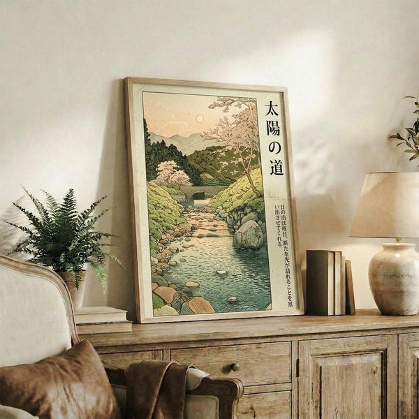May include: A framed Japanese woodblock-style poster displaying a serene mountain landscape with flowing stream, cherry blossoms, and traditional text characters, positioned on a rustic wooden sideboard alongside ferns in a vase, books, and ceramic pottery in a cozy living room with neutral tones and natural lighting.