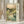 May include: Framed Japanese woodblock-style print showing a serene mountain landscape with cherry blossoms, a winding stream with stones, rolling hills, and traditional bridge. Features soft green and peach tones with Japanese text on cream background. Light wooden frame displayed on white surface against neutral curtained background with decorative dried wheat stems visible to the left.