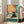 May include: Framed art print displayed in bright living room showing a black cat wearing yellow sunglasses lounging by a turquoise swimming pool. The vintage-style illustration features coral pink and teal walls, a large yellow sun, and a tropical drink with straw nearby. White frame leans against wall next to beige sofa.