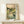 May include: Framed Japanese woodblock-style print displayed on light wood console table against white wall. Artwork depicts serene landscape with flowing stream, cherry blossom trees, stone bridge, rolling green hills and pale sun. Features traditional color palette of soft greens, browns and peachy tones with Japanese text on cream background. Small white wrapped gift box sits in front of frame.
