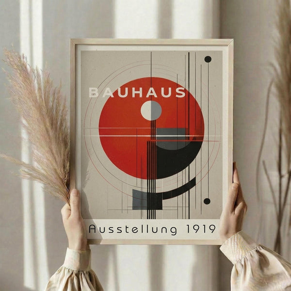 May include: Person holding framed geometric art print featuring red and black circles with linear elements on beige background, displayed in light wood frame. Dried wheat stalks visible on left side. Soft natural lighting creates warm, minimalist home decor scene with neutral tones throughout.