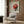 May include: Framed Bauhaus poster with red and black geometric circles and lines hung on wood-paneled wall above cream upholstered armchair with geometric pillow. Modern brass floor lamp and dark round side table with open book complete the mid-century modern reading nook scene.