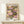 May include: Framed art print displaying vibrant water lilies in pink, purple, yellow and white blooms floating on green lily pads against dark water. Text reads "YAYOI KUSAMA" at top and "Modern Art 1984" at bottom. Light wood frame rests on pale wooden shelf beside small white wrapped gift with evergreen sprig. Soft shadows cast on cream wall background.