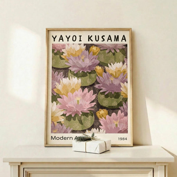 May include: Framed art print displaying vibrant water lilies in pink, purple, yellow and white blooms floating on green lily pads against dark water. Text reads "YAYOI KUSAMA" at top and "Modern Art 1984" at bottom. Light wood frame rests on pale wooden shelf beside small white wrapped gift with evergreen sprig. Soft shadows cast on cream wall background.