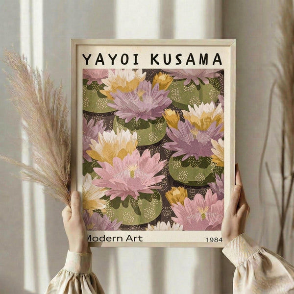 May include: Person holding framed poster featuring colorful water lilies in pink, yellow, and purple tones with green lily pads on dark water background. Light wood frame against cream curtains. Person wears beige ruffled sleeves and holds dried wheat stalks in other hand.