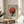 May include: Framed Bauhaus-style geometric poster with red and black circular design on beige background, displayed on round wooden side table next to beige upholstered chair. Natural sunlight creates soft shadows across white wall. Green plant leaves visible in corner. Wood frame complements minimalist modern interior styling.