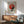 May include: Bauhaus-inspired geometric poster with red and black circular design on beige background, displayed in wooden frame above mid-century modern credenza. Green upholstered armchair with wooden arms sits nearby, white ceramic table lamp on credenza, natural sunlight casting shadows across white wall in minimalist living room setting.