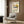 May include: Modern cream armchair with geometric pillow in contemporary room setting with wood paneled walls. Framed vintage-style poster showing colorful modernist chair design with yellow and blue accents against cream background. Gold floor lamp and dark round side table with open book complete the mid-century modern aesthetic.