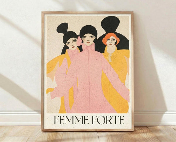 May include: A framed vintage-style poster featuring three stylized women with black hair silhouettes wearing flowing pink and yellow robes, with "FEMME FORTE" text below in black serif font. The artwork has an Art Deco aesthetic on cream background, displayed in a light wood frame leaning against a white wall on wooden flooring with soft natural lighting.