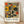 May include: Framed abstract art poster featuring geometric shapes in orange, yellow, and dark green on beige background, displayed in light wood frame against white wall on wooden floor with natural shadows