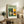 May include: Framed Bauhaus poster with modernist architectural illustration in green, yellow, and orange geometric shapes displayed on rustic wooden sideboard. Room setting shows neutral armchair, fern plant, ceramic vase, and stacked books creating warm, minimalist home decor scene with natural lighting.