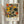 May include: Framed abstract art print displayed on white wall next to dried pampas grass stems. The artwork features geometric shapes and organic forms in orange, yellow, green, and beige tones arranged in a grid-like composition. Below the abstract design, black text reads "PAUL KLEE" with smaller text indicating "PLAYFUL ABSTRACTION" and "MUSEUM EDITION". The print has a light wooden frame and soft natural lighting creates gentle shadows on the white wall.