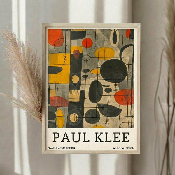 May include: Framed abstract art print displayed on white wall next to dried pampas grass stems. The artwork features geometric shapes and organic forms in orange, yellow, green, and beige tones arranged in a grid-like composition. Below the abstract design, black text reads "PAUL KLEE" with smaller text indicating "PLAYFUL ABSTRACTION" and "MUSEUM EDITION". The print has a light wooden frame and soft natural lighting creates gentle shadows on the white wall.