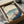 May include: Japanese woodblock-style print showing Mount Fuji with snow-capped peak, traditional buildings nestled among autumn trees with orange foliage, and turbulent blue waves in foreground. Artwork features muted teal, cream and coral tones with vertical Japanese text on left side. Print is displayed in black frame inside cardboard shipping box with protective tissue paper and wooden dowels for packaging.