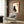 May include: Framed poster with Japanese calligraphy and black cat silhouette displayed on wood-paneled wall above beige upholstered chair with geometric pillow. Poster features orange border, cream background, sitting black cat, and two kanji characters. Modern living room setting with brass floor lamp and side table.