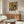 May include: Framed Paul Klee abstract art poster with colorful geometric shapes in yellow, orange, green, and red displayed above a wooden sideboard in a modern living room. White curved chair and glass coffee table visible in foreground. Clean minimalist interior with white walls and natural lighting.