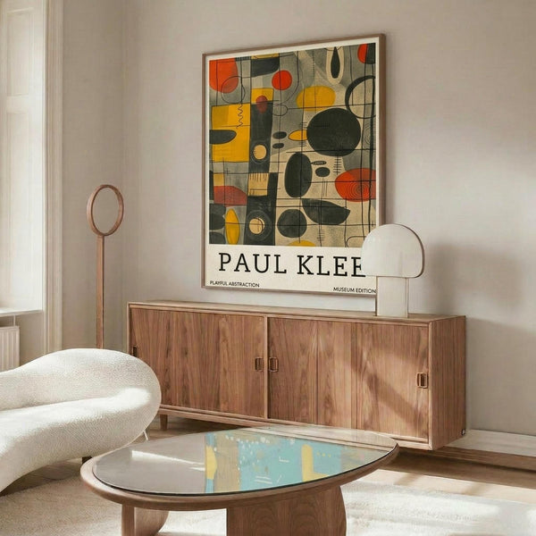 May include: Framed Paul Klee abstract art poster with colorful geometric shapes in yellow, orange, green, and red displayed above a wooden sideboard in a modern living room. White curved chair and glass coffee table visible in foreground. Clean minimalist interior with white walls and natural lighting.