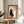 May include: Framed poster featuring a black cat silhouette sitting upright with Japanese characters, displayed on a round wooden side table next to a beige upholstered chair. The artwork has an orange-brown mat border within a light wooden frame, set against white walls with natural sunlight streaming across the scene and a plant visible in the corner.